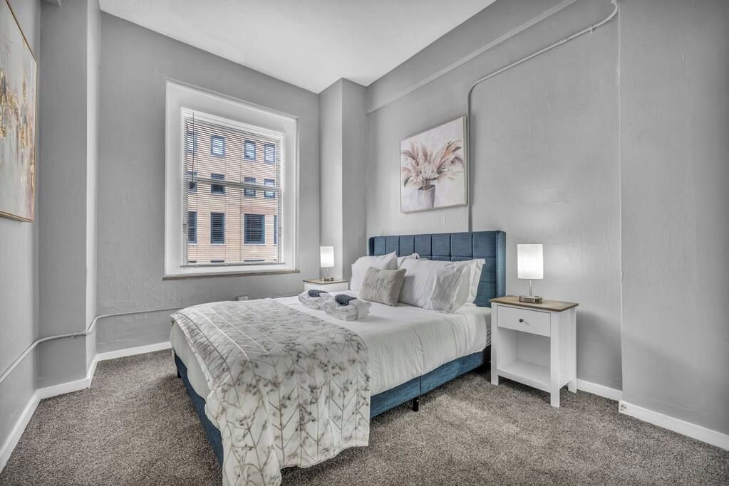 Un dormitorio con cama, mesitas de noche, ventana y un cuadro en la pared.