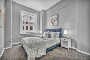 Un dormitorio con cama, mesitas de noche, ventana y un cuadro en la pared.
