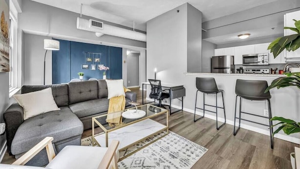 A modern living room with a grey sofa, a coffee table, and a small desk with a chair. A kitchen area with a microwave and a sink is visible in the background. - Modern 1B in Heart of Downtown • Pool & Spa • Gym (Cleveland)