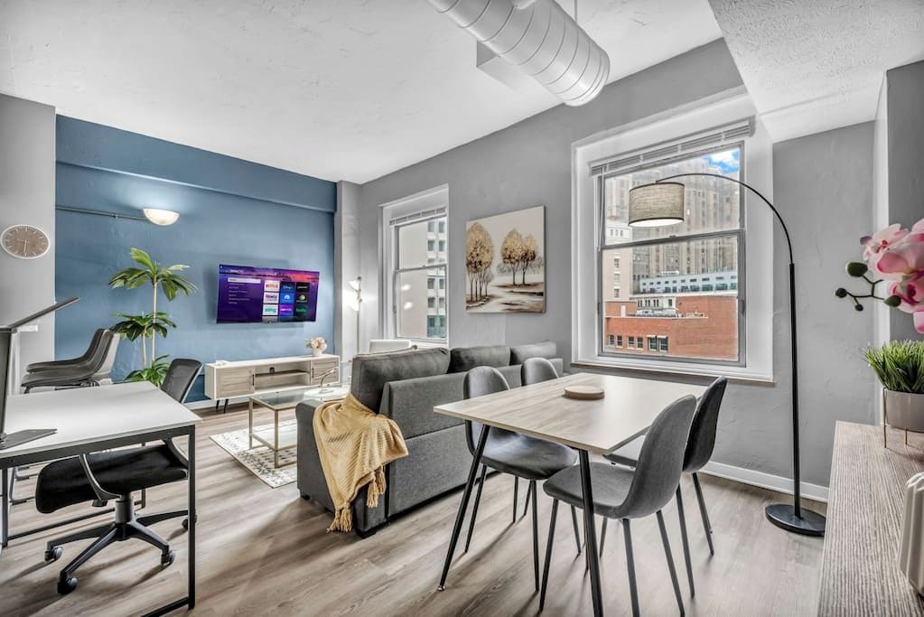 A modern office space with a gray sofa, a dining table with chairs, a conference table with chairs, a large window with a city view, and a mounted TV displaying a program.