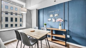 A modern dining area with a wooden table and grey chairs, a blue accent wall, and a window overlooking a building. - Modern 1B in Heart of Downtown • Pool & Spa • Gym (Cleveland)