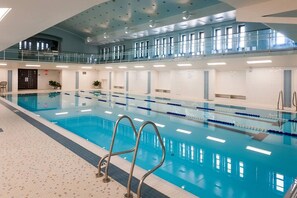 An indoor swimming pool with clear lanes, a mezzanine level with windows, and a tiled floor.