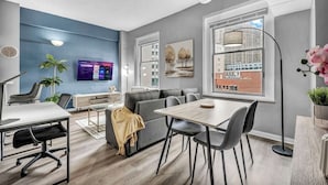A modern office space with a gray sofa, a dining table with chairs, a conference table with chairs, a large window with a city view, and a mounted TV displaying a program. - Modern 1B in Heart of Downtown • Pool & Spa • Gym (Cleveland)