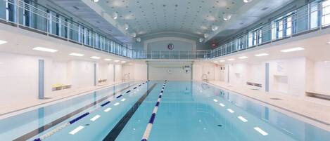 Una amplia piscina interior con carriles bien definidos, techo alto con luces distribuidas uniformemente y barandilla de vidrio en el nivel superior.