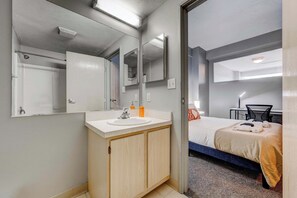 A hotel room with a bathroom featuring a sink, mirror, and shower. The room has a bed with a blanket and pillows, a desk with a chair, and a small table with a lamp. - HUGE Loft in Heart of Downtown • Pool/Sauna • Gym (Cleveland)