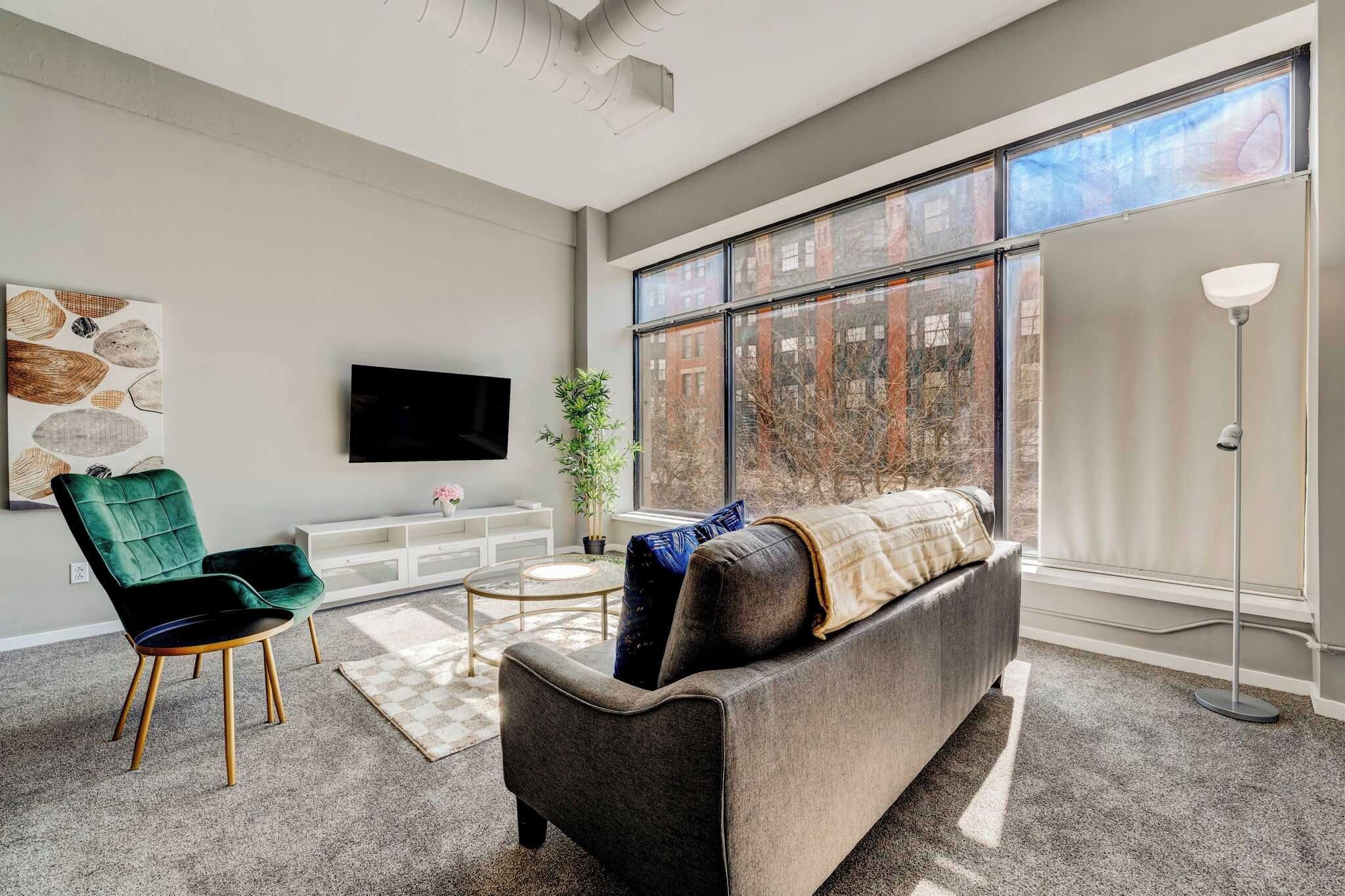 A modern living room with a grey sofa, a green armchair, and a flat-screen TV mounted on the wall.