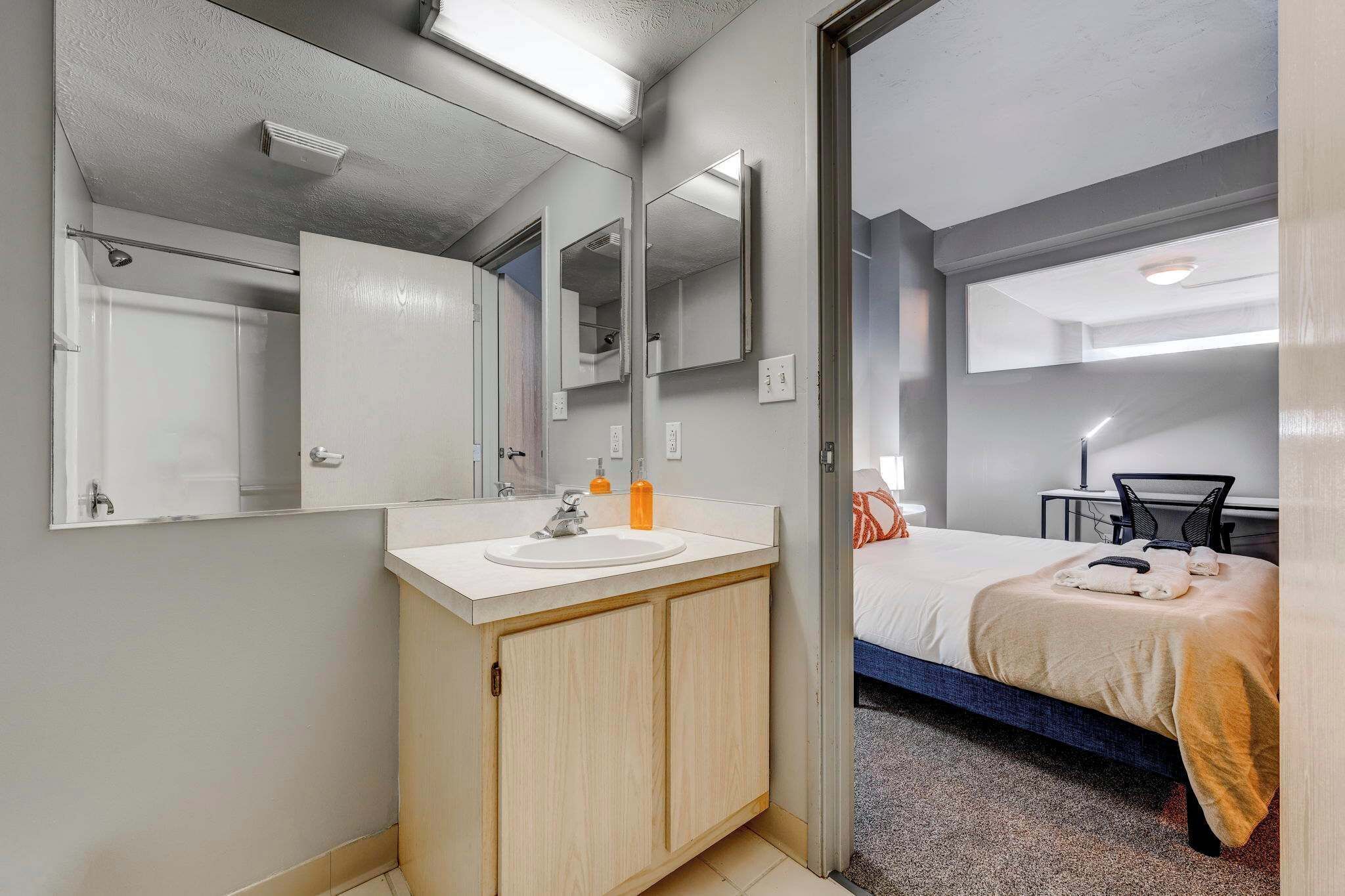 A hotel room with a bathroom featuring a sink, mirror, and shower. The room has a bed with a blanket and pillows, a desk with a chair, and a small table with a lamp.