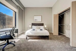 A hotel room with a large bed, a desk with a chair, a window with a view of buildings, and a closet with a hanging rack. - HUGE Loft in Heart of Downtown • Pool/Sauna • Gym (Cleveland)