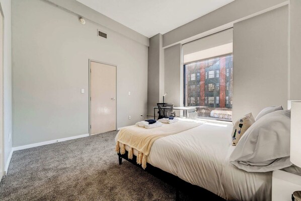 A modern bedroom with a large window, a bed with a beige blanket, and a small table with a chair. - HUGE Loft in Heart of Downtown • Pool/Sauna • Gym (Cleveland)