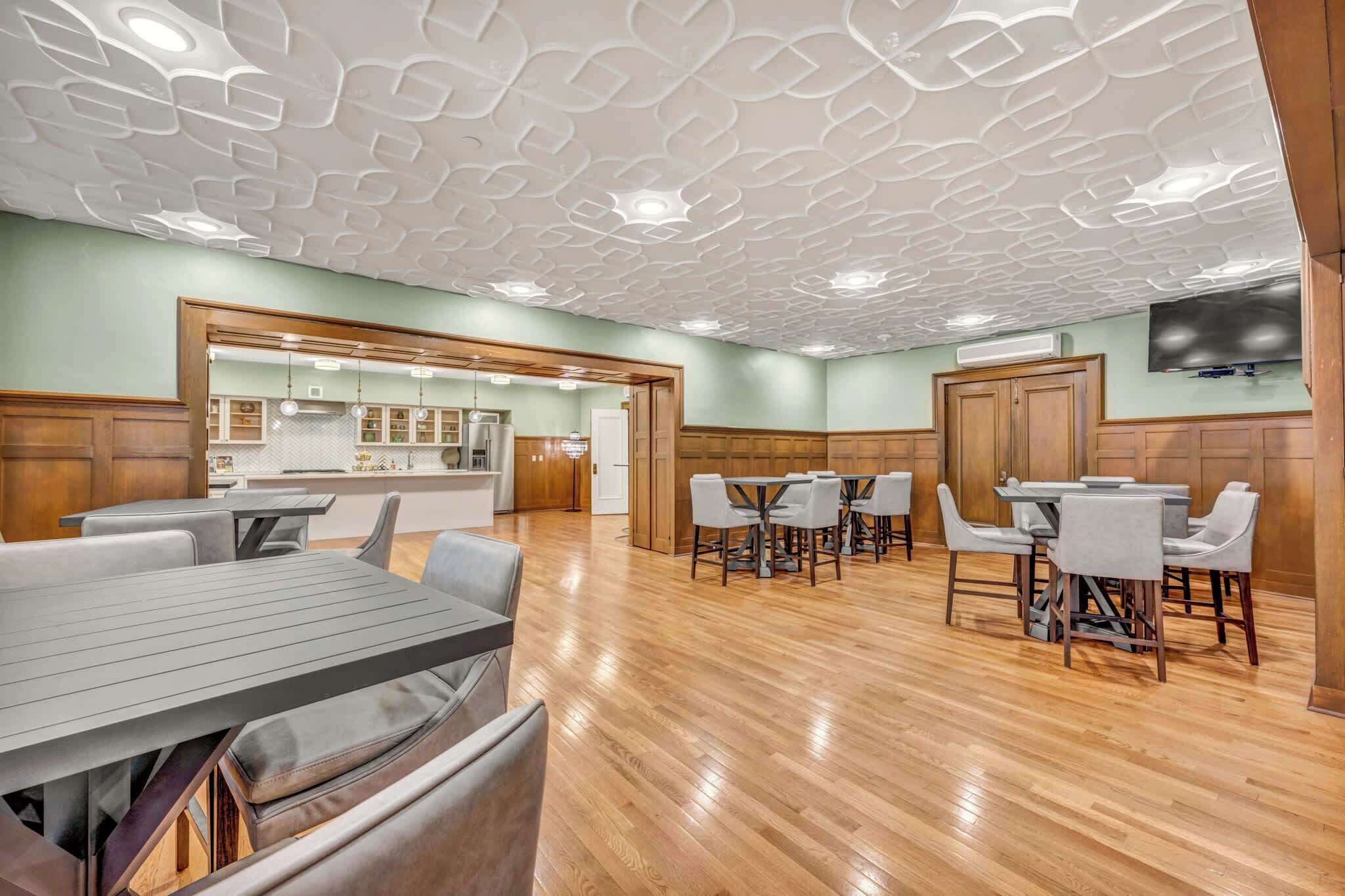 A spacious dining area with wooden flooring, a modern ceiling design, and a kitchen visible in the background.