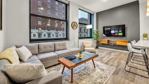 A modern living room with a large window, a sofa, a coffee table, a TV, and a small dining area. - Central Flat w/ Pool & Sauna • Gym•Heart of DT (Cleveland)