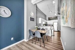 A modern kitchen with white cabinets, a dining area with a wooden table and grey chairs, and a wall-mounted clock.