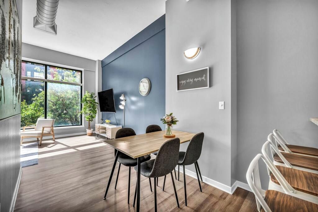 A modern dining area with a wooden table and chairs, a large window with a view of greenery, and a wall-mounted clock.