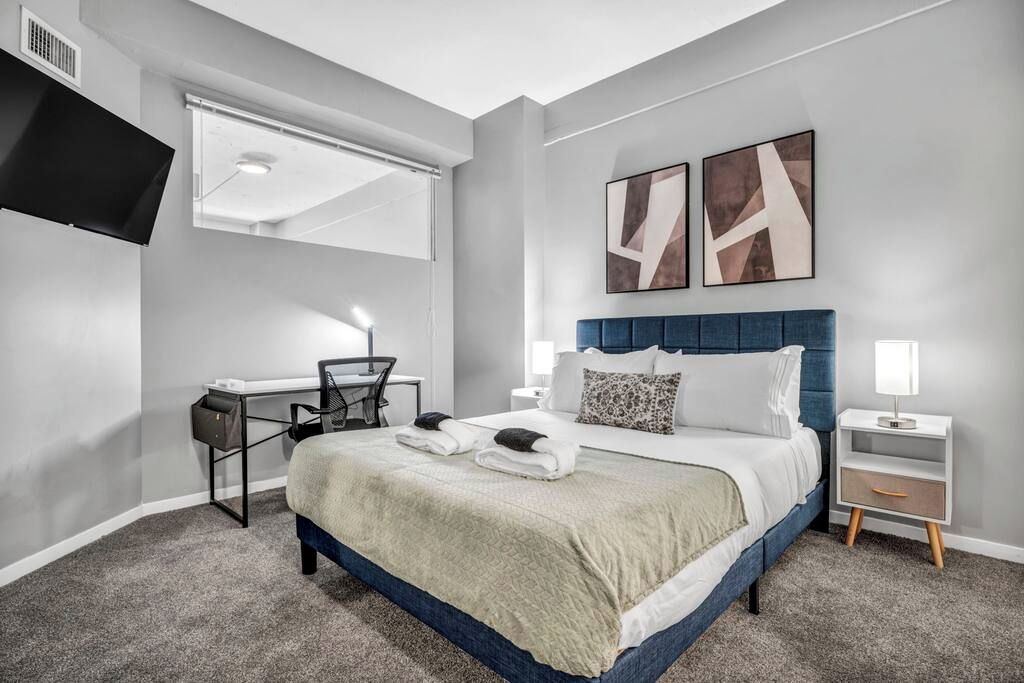 A modern hotel room with a large bed, a desk, a chair, a TV, and artwork on the wall.