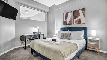 A modern hotel room with a large bed, a desk, a chair, a TV, and artwork on the wall.