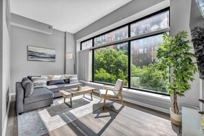 Un salon moderne avec un canapé gris, une table basse et un fauteuil. Une grande fenêtre offre une vue sur les arbres et les bâtiments.