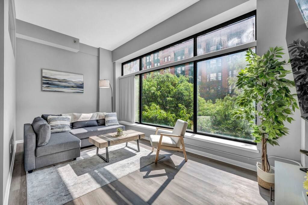 A modern living room with a grey sofa, a coffee table, and a chair. There is a large window with a view of trees and buildings.