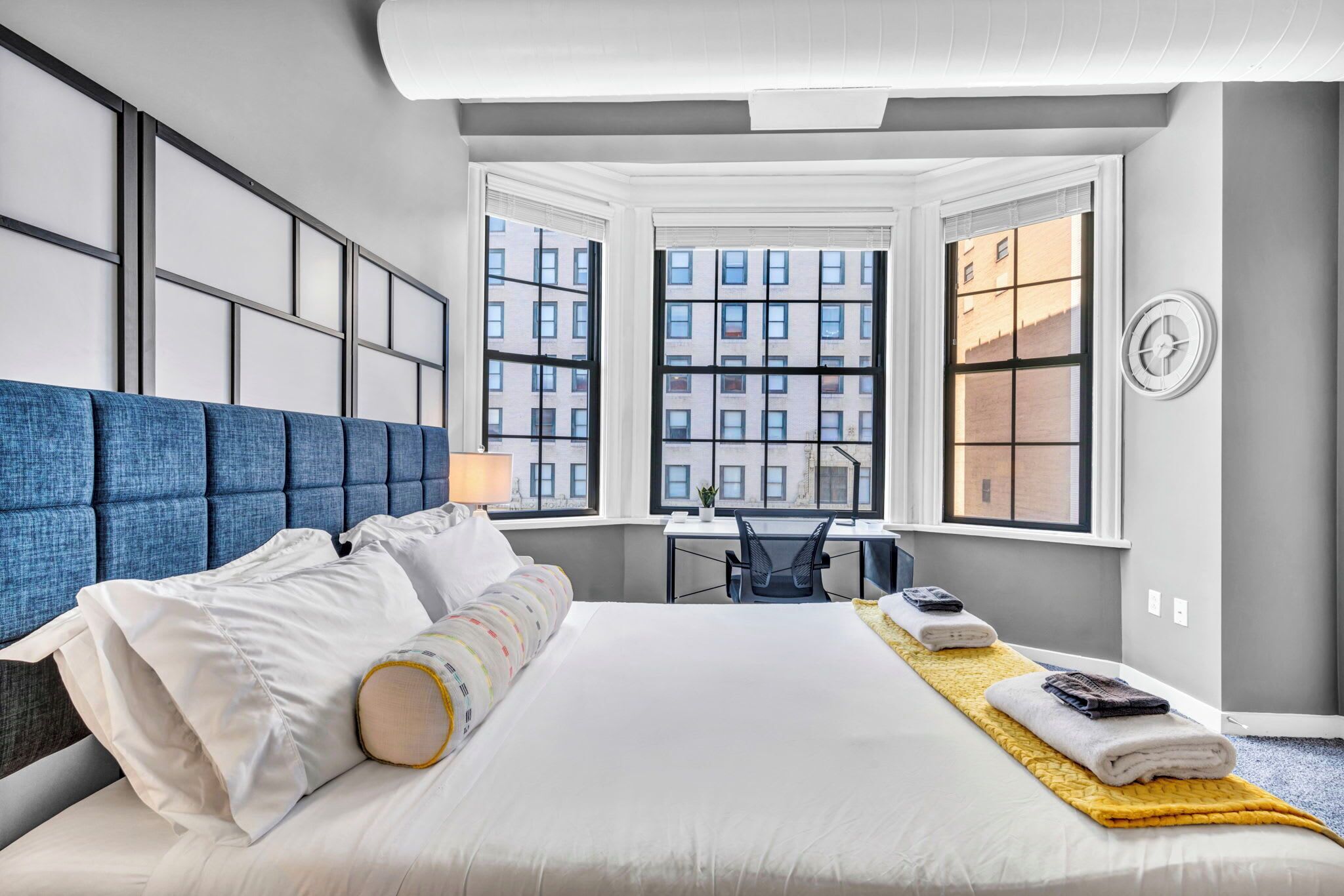 Habitación de hotel con una cama grande, un escritorio y vistas a edificios a través de las ventanas.