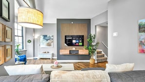 Un salon moderne avec un téléviseur à écran plat fixé sur un mur recouvert de panneaux en bois, un grand luminaire suspendu et un escalier menant à un niveau supérieur.