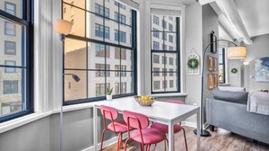 Un salon moderne avec une table blanche, des chaises rouges, un canapé gris et de grandes fenêtres donnant sur des immeubles.