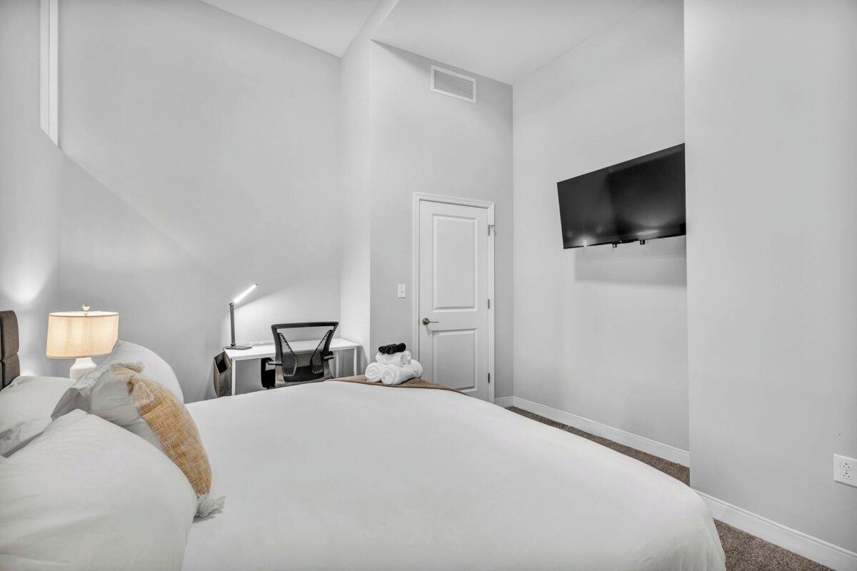 Habitación de hotel con una cama grande, un escritorio con lámpara, una silla, un televisor montado en la pared y una puerta.