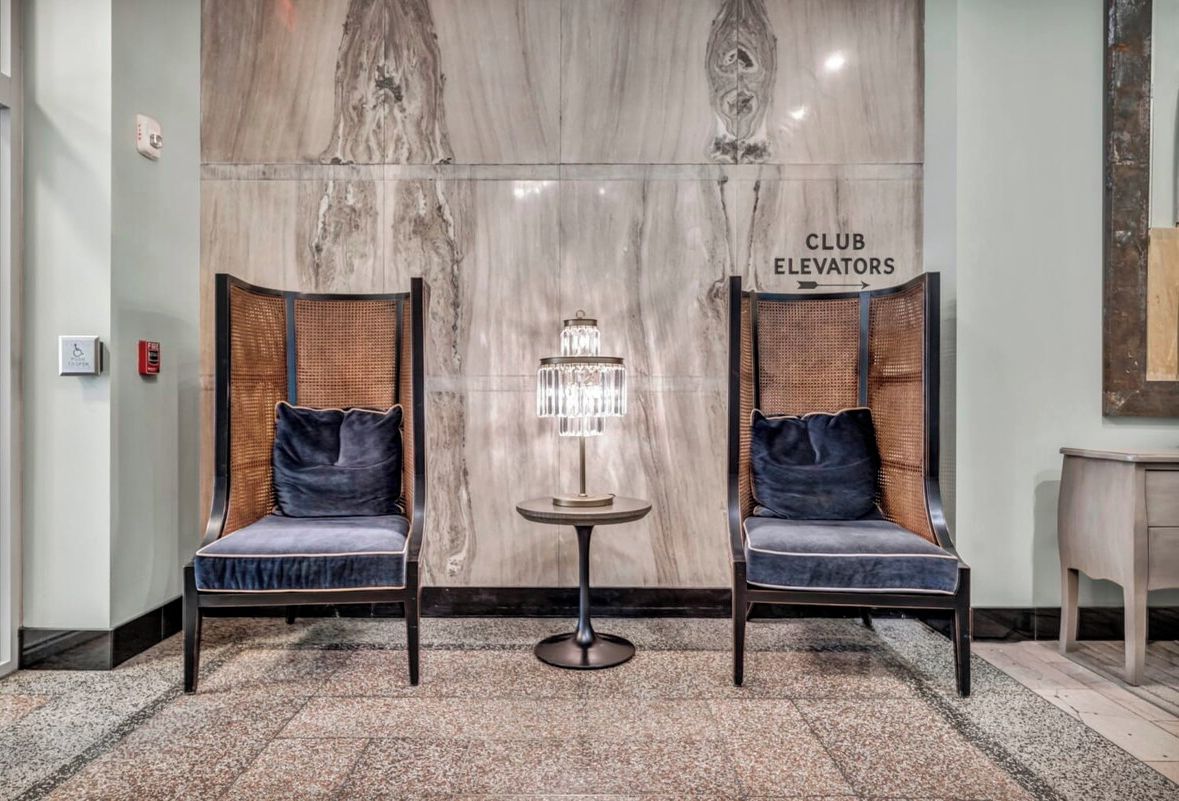 Eine Lobby mit zwei Polsterstühlen, einem kleinen Tisch mit einer Kristalllampe und einer Marmorwand mit der Aufschrift 'CLUB ELEVATORS'.
