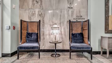 Un vestíbulo con dos sillones tapizados, una mesita con lámpara de cristal y una pared de mármol con el letrero 'CLUB ELEVATORS'.