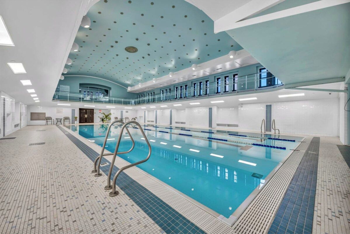 A large indoor swimming pool with a tiled floor, a blue ceiling with circular patterns, and a second level balcony with windows.