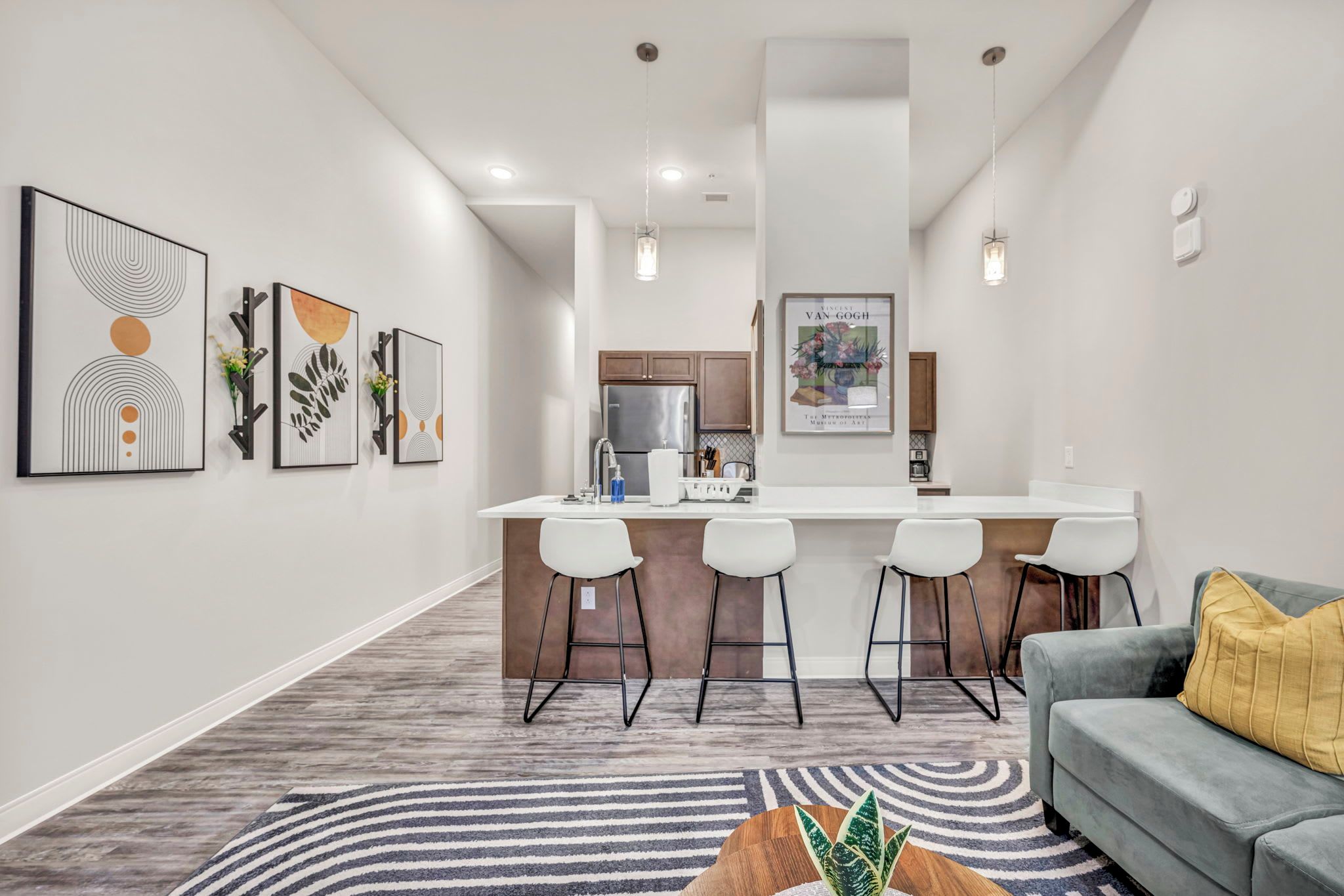 Una cocina moderna con una isla central, taburetes de bar y un área de comedor con sofá gris y alfombra estampada.