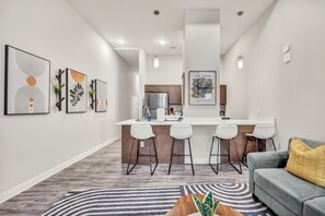 A modern kitchen with a central island, bar stools, and a dining area with a gray sofa and a patterned rug.