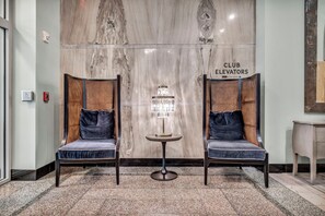A lobby with two upholstered chairs, a floor lamp, and a wall with a sign reading 'CLUB ELEVATORS'.