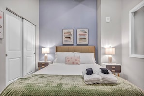 A neatly arranged bedroom with a bed, two bedside tables, a nightstand, a green quilted bedspread, two wall-mounted picture frames, and a white door. - Central 1B in Heart of Downtown • Pool & Spa • Gym (Cleveland)