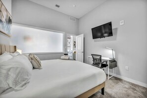 A modern hotel room with a large bed, a desk with a computer, a flat-screen TV, and a window with a view of the hallway. - Central Flat w/ Pool & Sauna • Gym•Heart of DT (Cleveland)