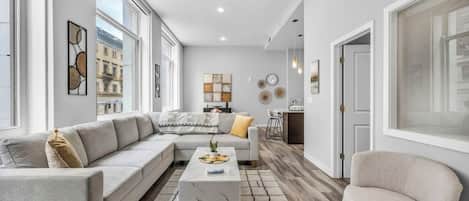 Una sala de estar moderna con un sofá seccional, una mesa de centro y una silla. Hay ventanas amplias, obras de arte en las paredes y una cocina visible al fondo.