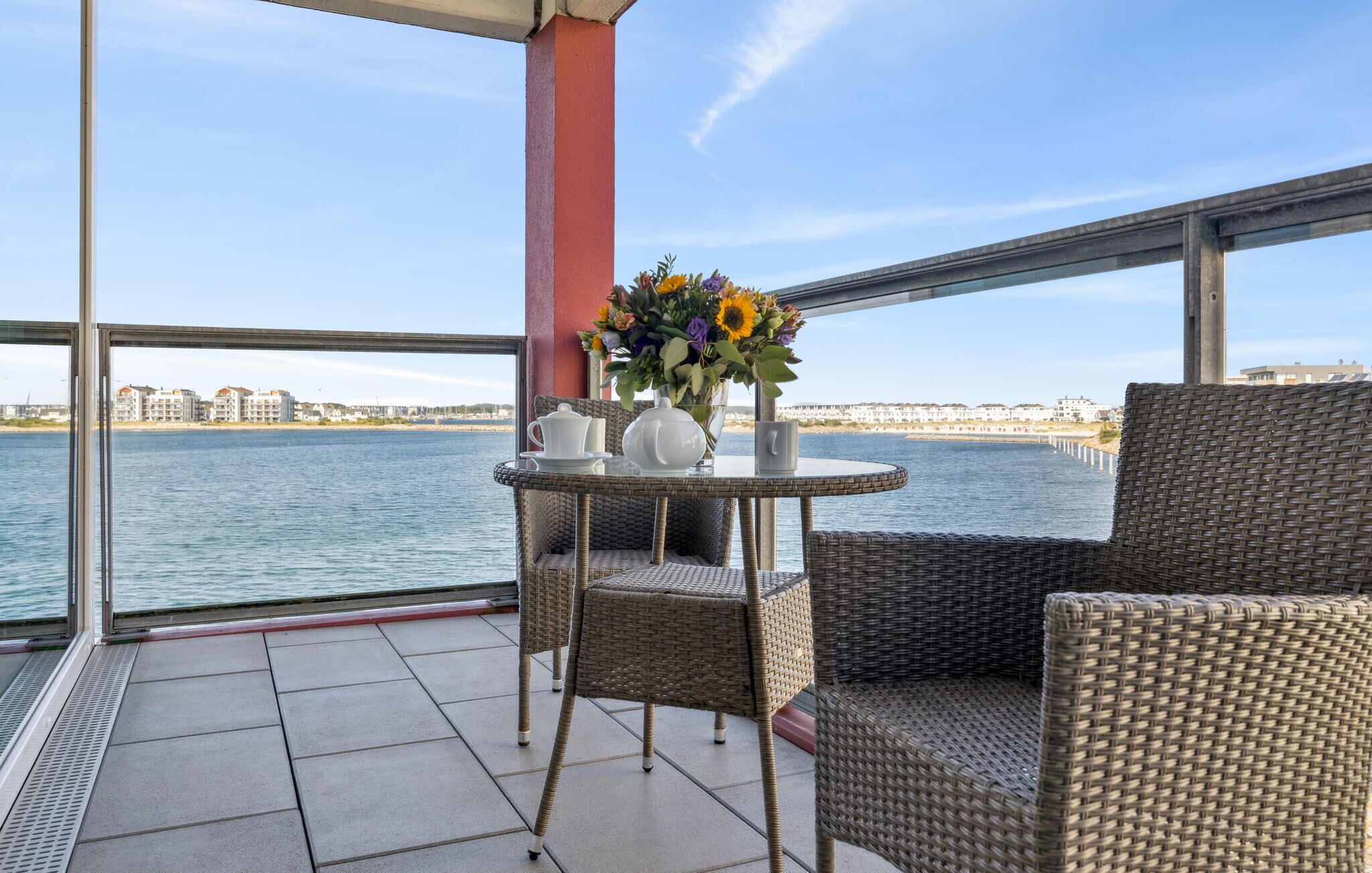 A balcony with wicker furniture, a table with a vase of flowers, and a view of a waterfront with buildings.