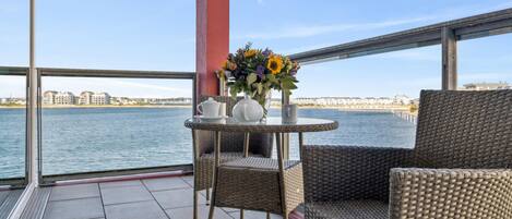 A balcony with wicker furniture, a table with a vase of flowers, and a view of a waterfront with buildings.
