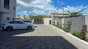 A paved driveway leading to a gated entrance, a white car parked on the left, and a building with multiple windows and balconies in the background. - Enjoyable apartment with AC in Kingston (Kingston)
