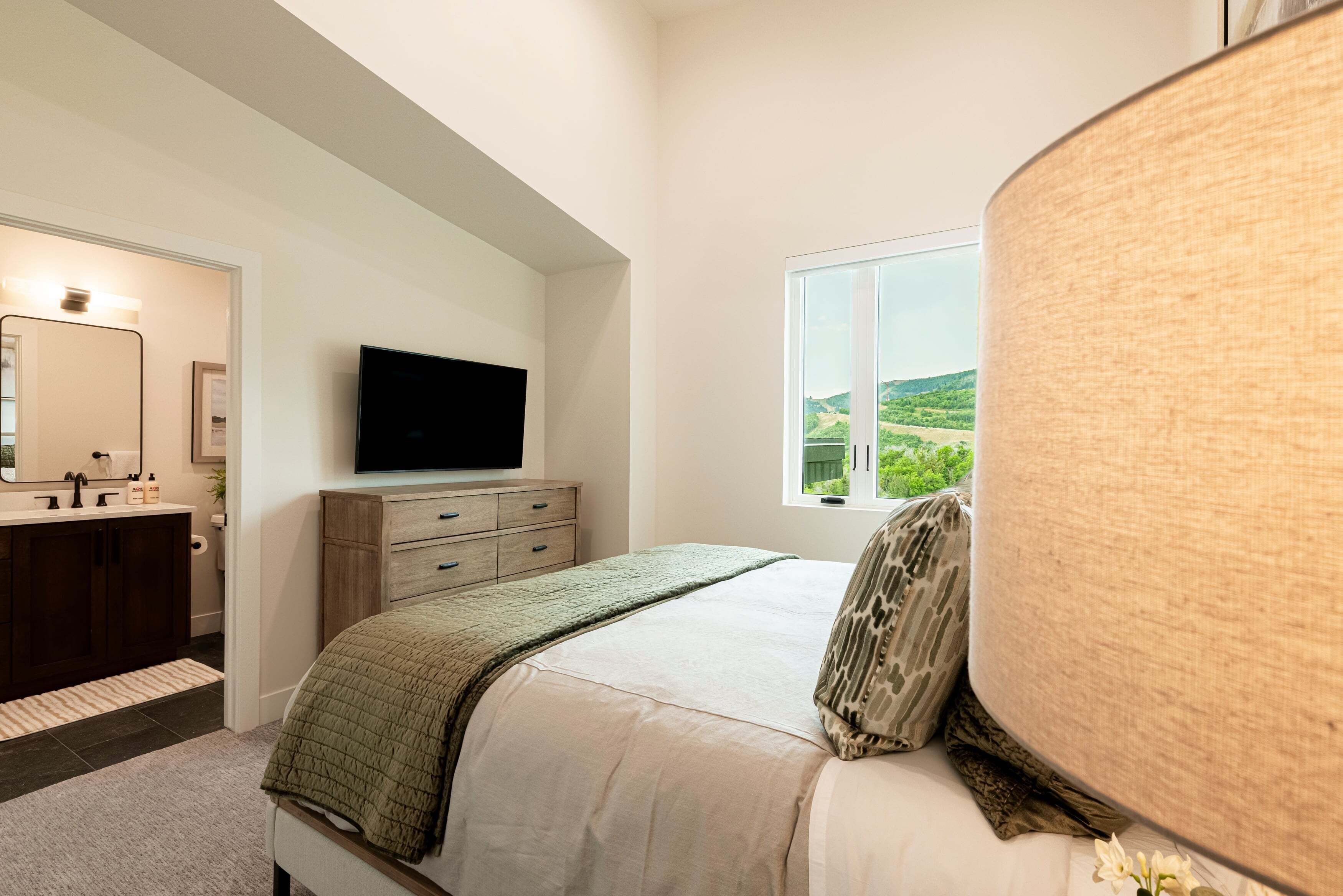 Un dormitorio con cama, televisor, ventana con vistas y baño con lavamanos y espejo.