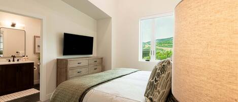 Un dormitorio con cama, televisor, ventana con vistas y baño con lavamanos y espejo.