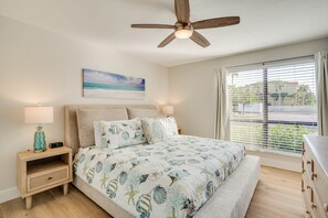 Ein Schlafzimmer mit Bett, Nachttischen, Deckenventilator, Fenster mit Jalousien und einem Wandbild zum Strandthema.