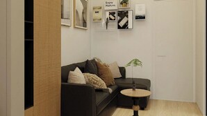 Un salon moderne avec un canapé gris foncé, une petite table basse ronde et une plante en pot. Des tableaux encadrés ornent le mur, et une porte est visible au fond.