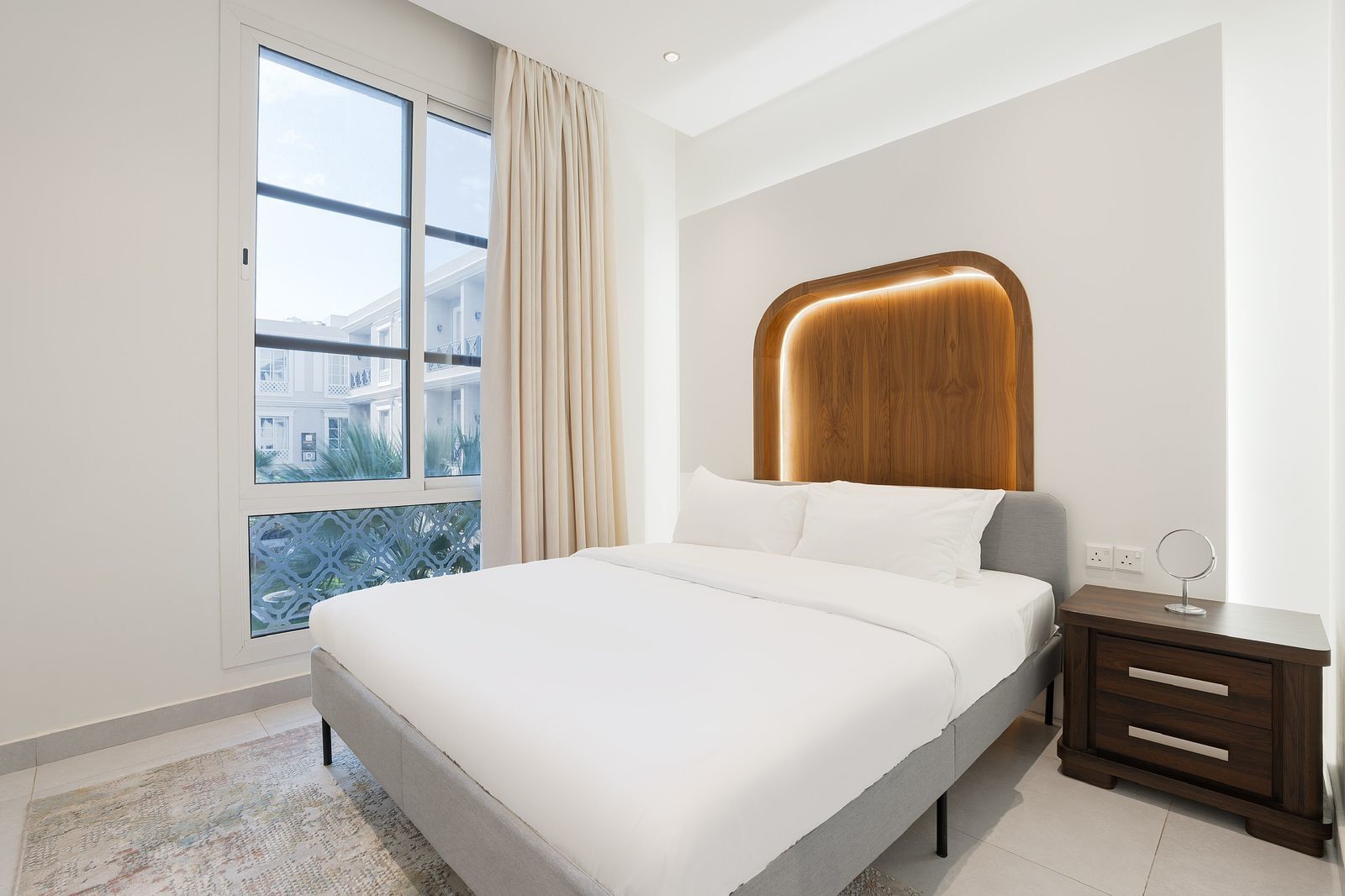 A modern bedroom with a large bed, a wooden headboard, a bedside table with a mirror, and a view of a building through a window.