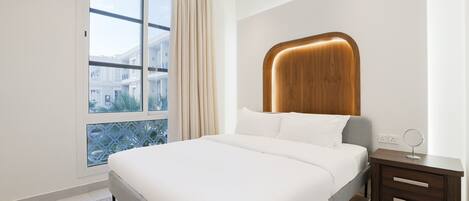 Un dormitorio moderno con una cama grande, cabecera de madera, mesita de noche con espejo y vista a un edificio a través de una ventana.