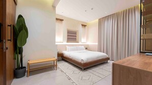 A modern bedroom with a bed, bedside table, bench, potted plant, and a large window with curtains. - Luxury Apartment in Alwadi (Riyadh)