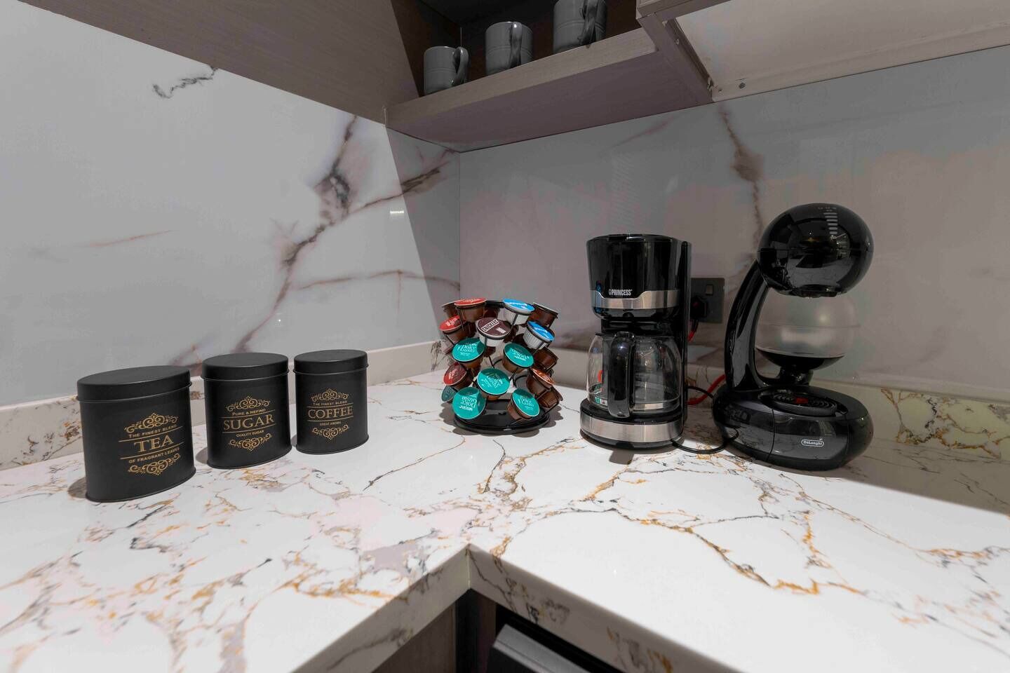 A kitchen counter with three black containers labeled 'TEA', 'SUGAR', and 'COFFEE', a coffee maker, and a coffee pod holder.