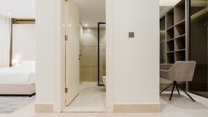 A modern hotel room with a bed, a desk, and a chair. There is a bathroom with a shower and toilet. The room has a large window and a closet with shelves. - Three BR Spacious & Luxurious Flat in Hittin (Riyadh)