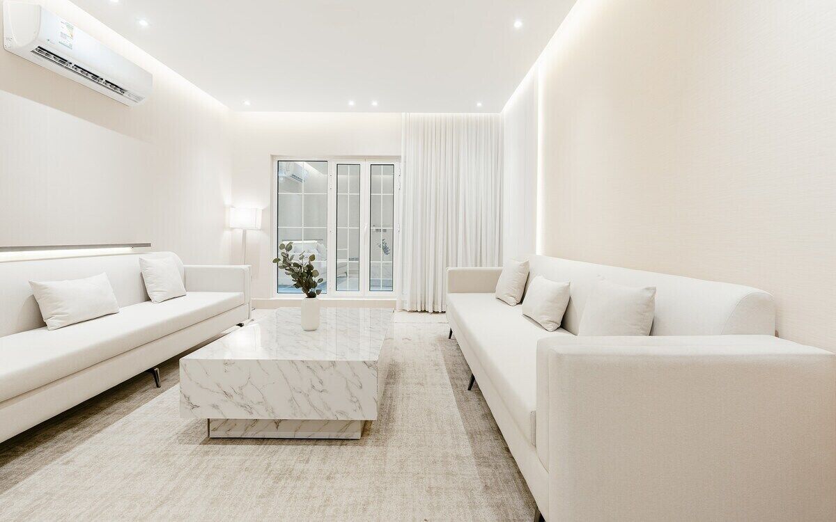 A modern, minimalist living room with a white sofa, a marble coffee table, and a view of the outdoors through large glass doors.