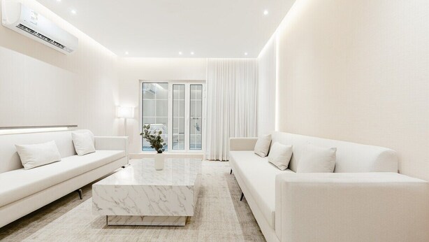 A modern, minimalist living room with a white sofa, a marble coffee table, and a view of the outdoors through large glass doors. - Three BR Spacious & Luxurious Flat in Hittin (Riyadh)