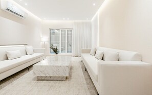 Una sala de estar moderna y minimalista con un sofá blanco, una mesa de centro de mármol y vistas al exterior a través de grandes puertas de vidrio.