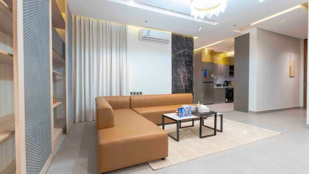 A modern living room with a sectional sofa, a coffee table, and a kitchen area in the background. - Apartment Luxurious and Modern Design in Alwadi Xl (Riyadh)